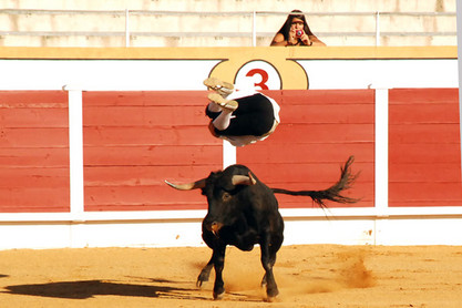 Mann springt in Arena über Stier, Frau auf Sitzbank fotografiert das